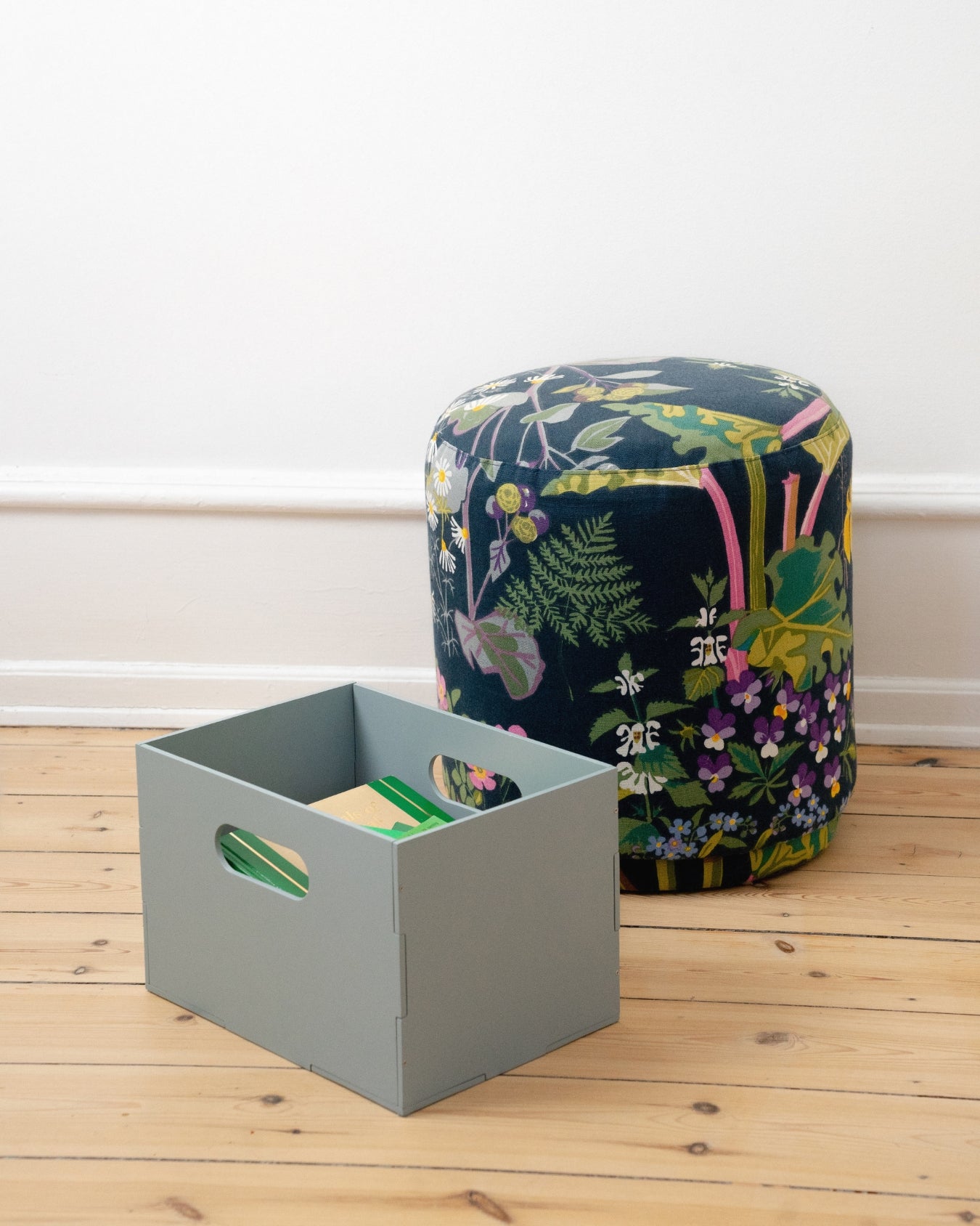 Gray storage box and floral-patterned ottoman on a wooden floor.