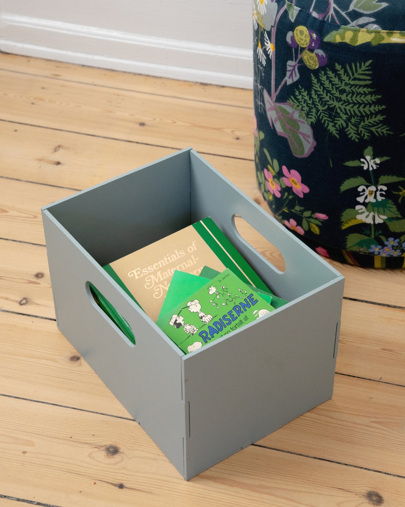 Gray storage box with green items on a wooden floor next to a floral-patterned bag.