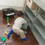 Child playing with a toy piano on a wooden floor next to a bookshelf.
