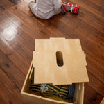 Child playing with toy cars on a wooden floor next to a wooden box.