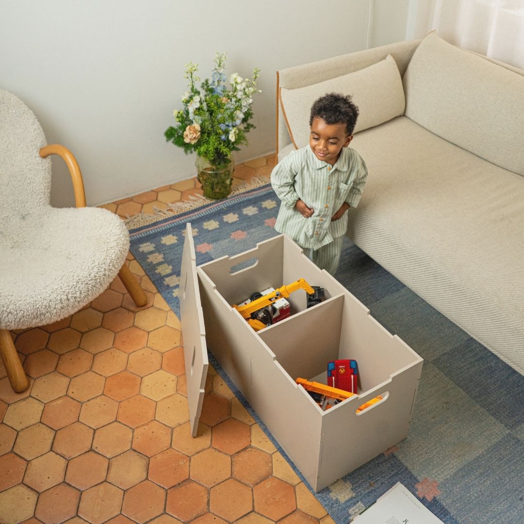 Child standing next to a storage bin filled with toys in a living room.