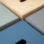 Close-up of four rectangular storage boxes or bins arranged in a corner pattern, showing different finish options: light natural wood, muted sage green, slate blue, and light gray. Each box has a recessed handle cutout on the top surface.