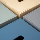 Close-up of four rectangular storage boxes or bins arranged in a corner pattern, showing different finish options: light natural wood, muted sage green, slate blue, and light gray. Each box has a recessed handle cutout on the top surface.