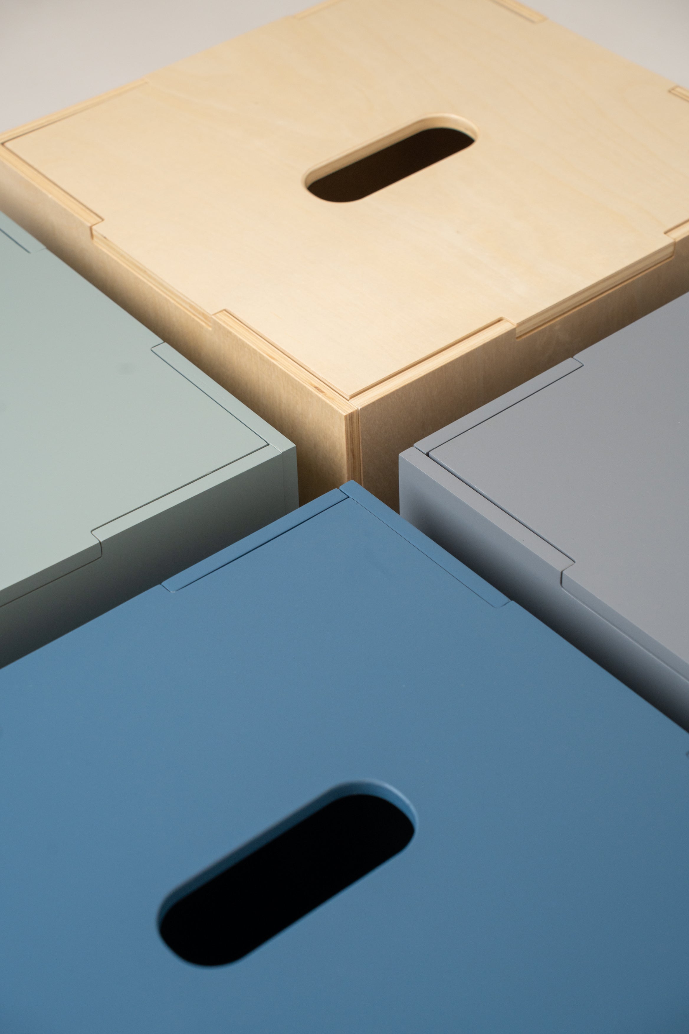 Close-up of four rectangular storage boxes or bins arranged in a corner pattern, showing different finish options: light natural wood, muted sage green, slate blue, and light gray. Each box has a recessed handle cutout on the top surface.