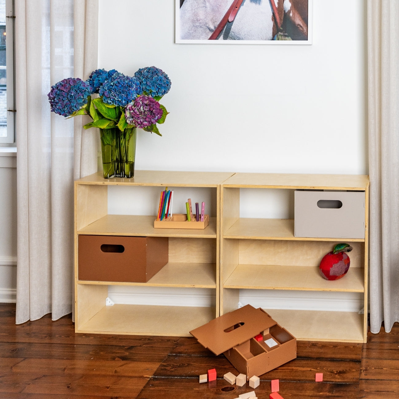 Wooden shelf with storage boxes and toys on a wooden floor, with a vase of flowers and a framed picture in the background.