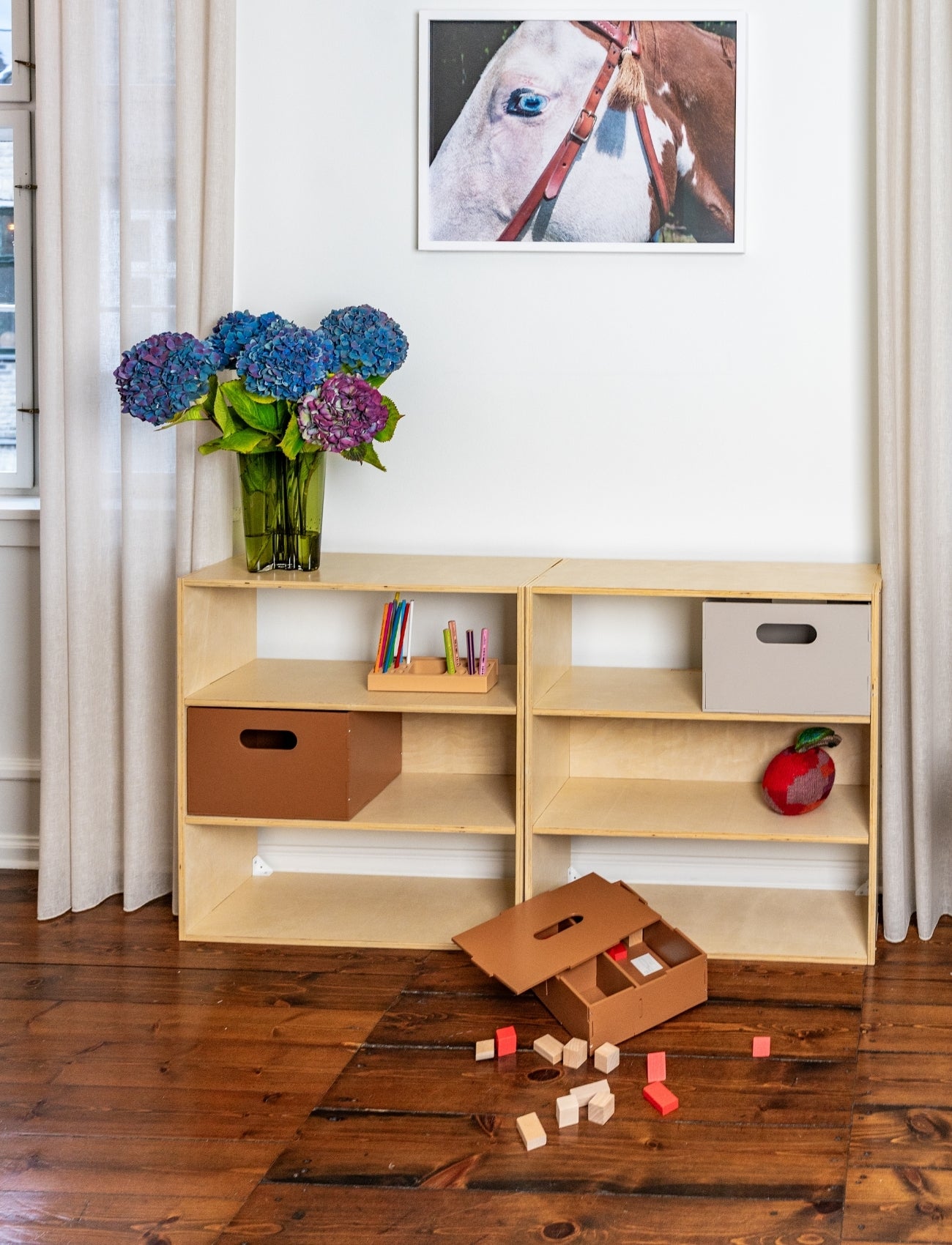 Wooden shelf with storage boxes and toys on a wooden floor, with a vase of flowers and a framed picture in the background.