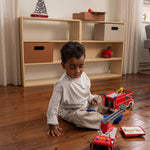 Child playing with toy fire trucks on a wooden floor in a room with a bookshelf.