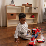 Child playing with toy fire trucks on a wooden floor in a room with a bookshelf.