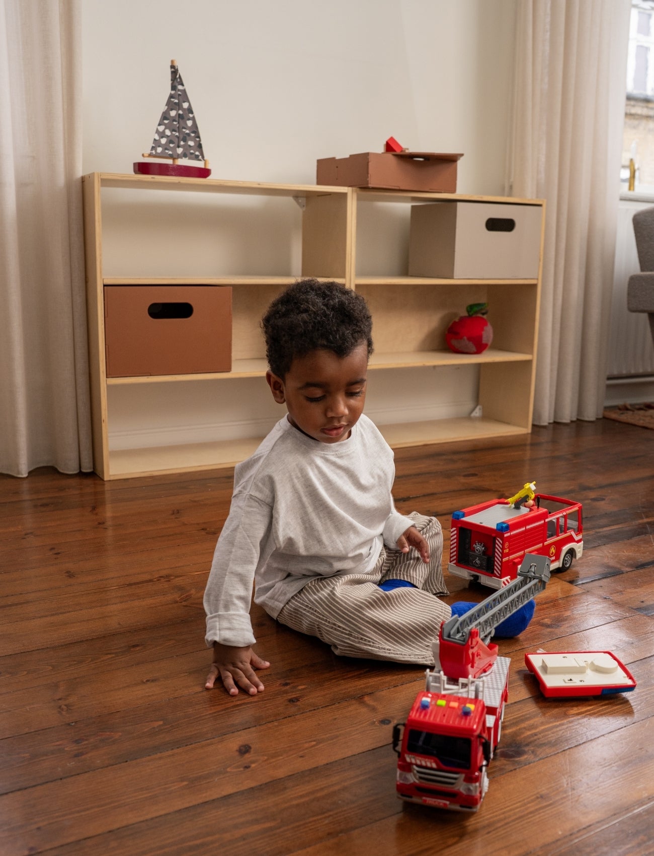 Child playing with toy fire trucks on a wooden floor in a room with a bookshelf.