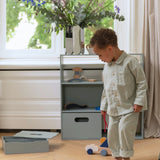 Child in striped pajamas standing next to a gray storage unit with toys in a room with a window and flowers.