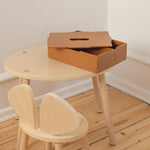 A small round light wood table with matching wooden chair and a brown cardboard storage box on top, photographed in a minimalist room with white walls and light wood flooring.