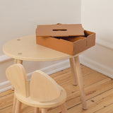 A small round light wood table with matching wooden chair and a brown cardboard storage box on top, photographed in a minimalist room with white walls and light wood flooring.