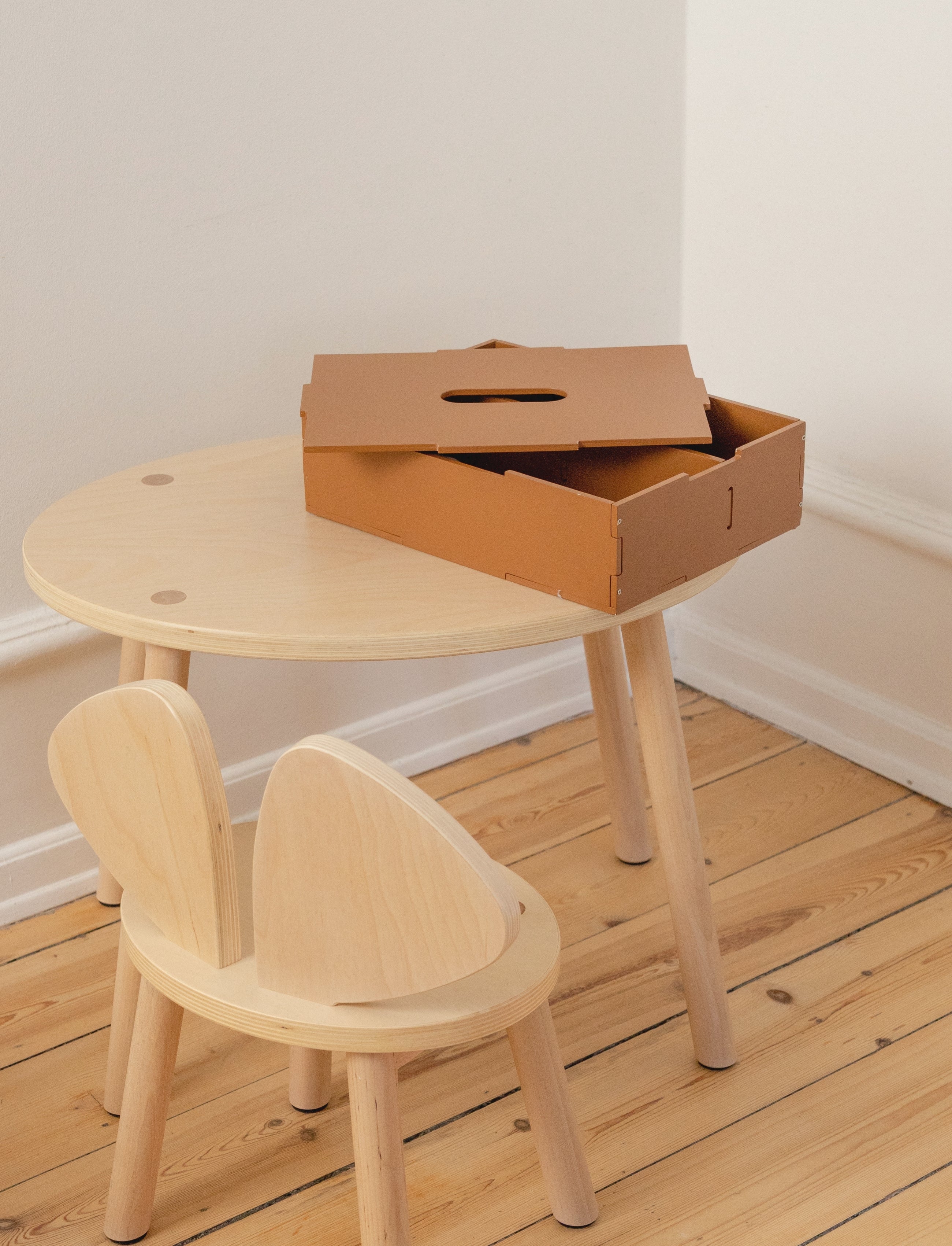 A small round light wood table with matching wooden chair and a brown cardboard storage box on top, photographed in a minimalist room with white walls and light wood flooring.
