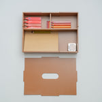 A minimalist wooden desk organizer mounted on a light gray wall, shown with its matching flat back panel below. The organizer contains pink and orange pencils, a yellow pencil, a beige folder, and a small white striped item.