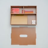 A minimalist wooden desk organizer mounted on a light gray wall, shown with its matching flat back panel below. The organizer contains pink and orange pencils, a yellow pencil, a beige folder, and a small white striped item.