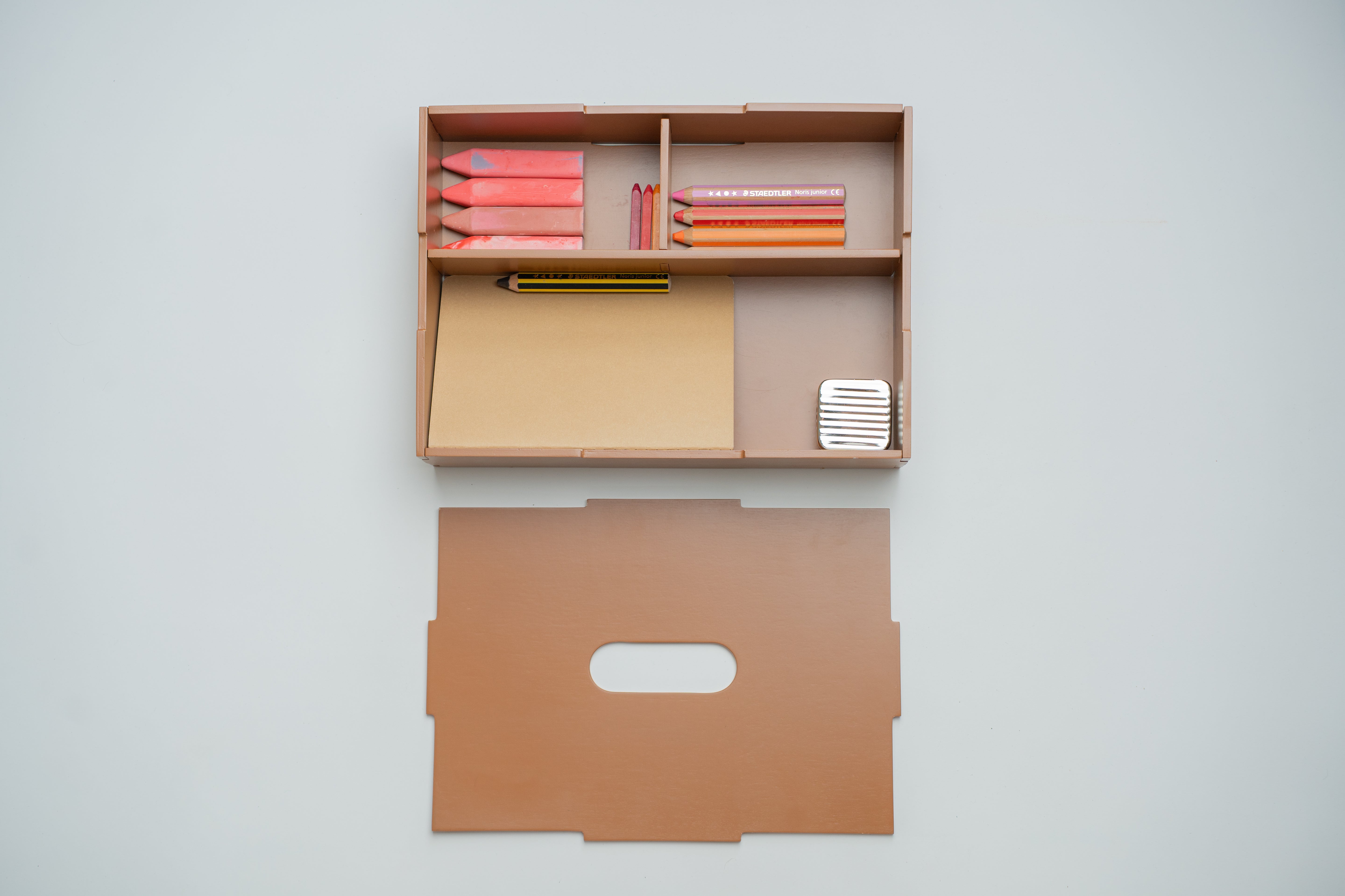 A minimalist wooden desk organizer mounted on a light gray wall, shown with its matching flat back panel below. The organizer contains pink and orange pencils, a yellow pencil, a beige folder, and a small white striped item.