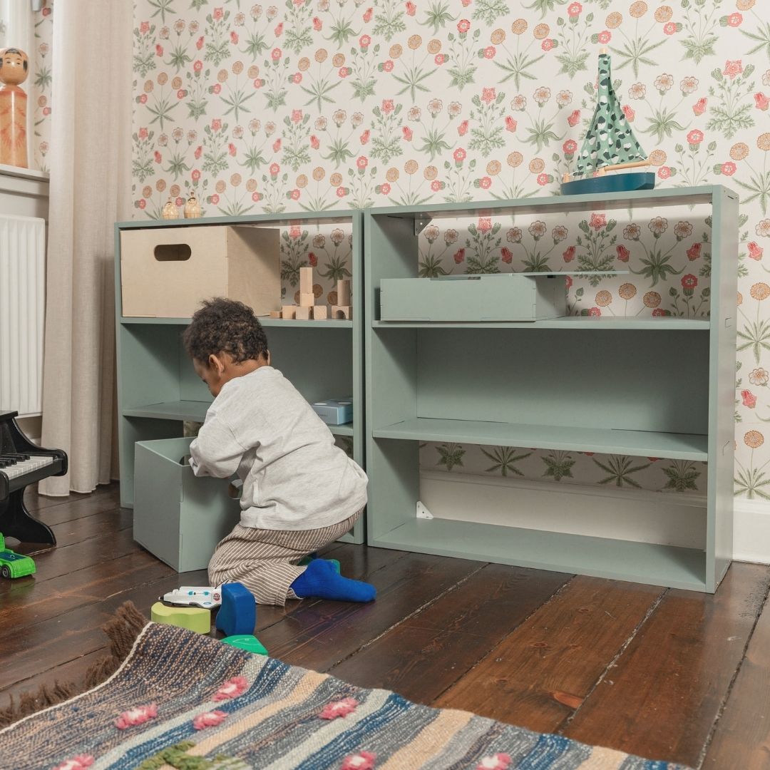 Child playing with a green toy cabinet in a room with floral wallpaper.