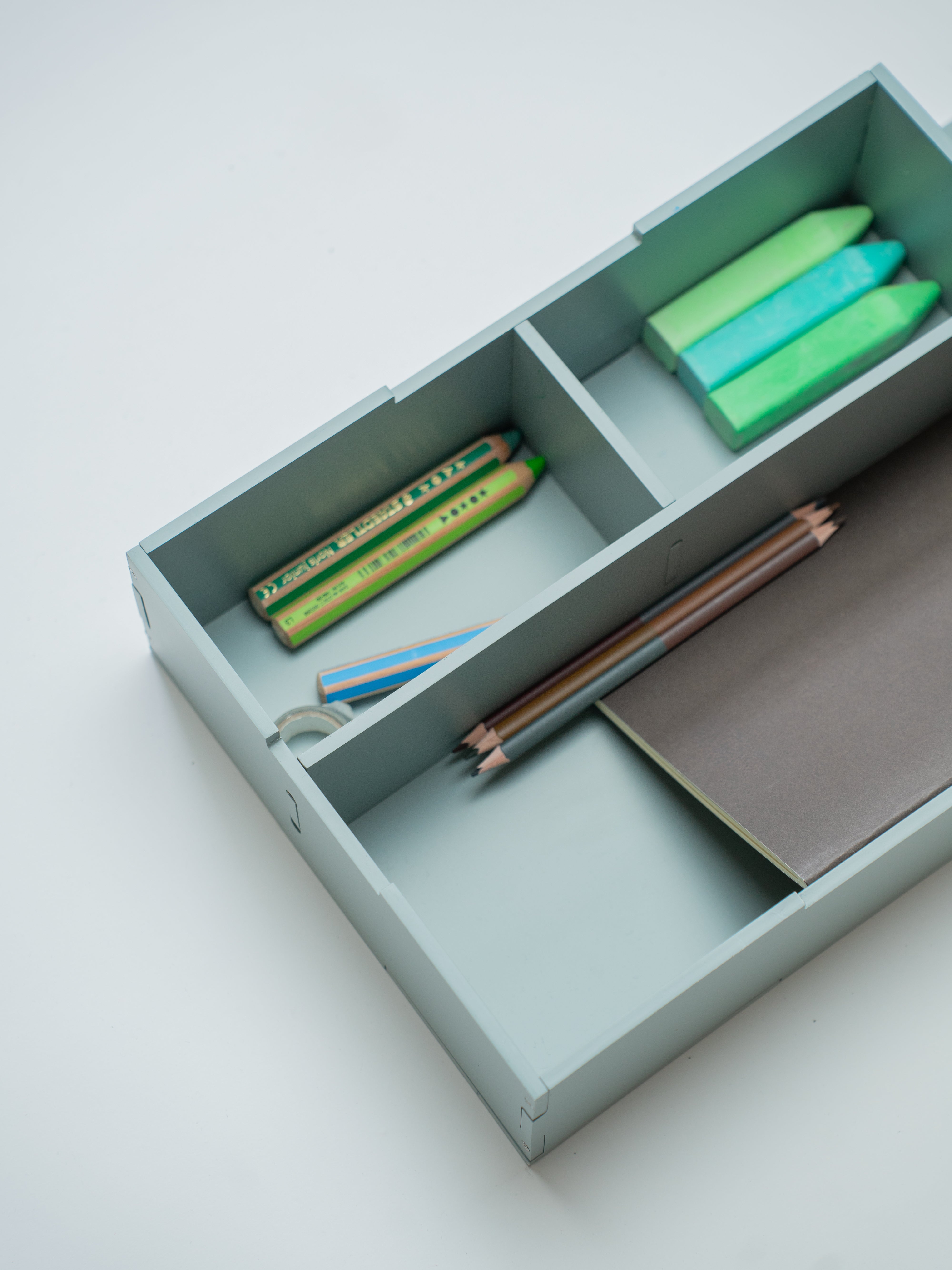 A light olive green wooden drawer organizer with three compartments, shown open and styled with colorful pencils, a ruler, and a notebook on a clean white background.
