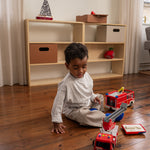 Child playing with toy fire trucks on a wooden floor in a room with shelves.