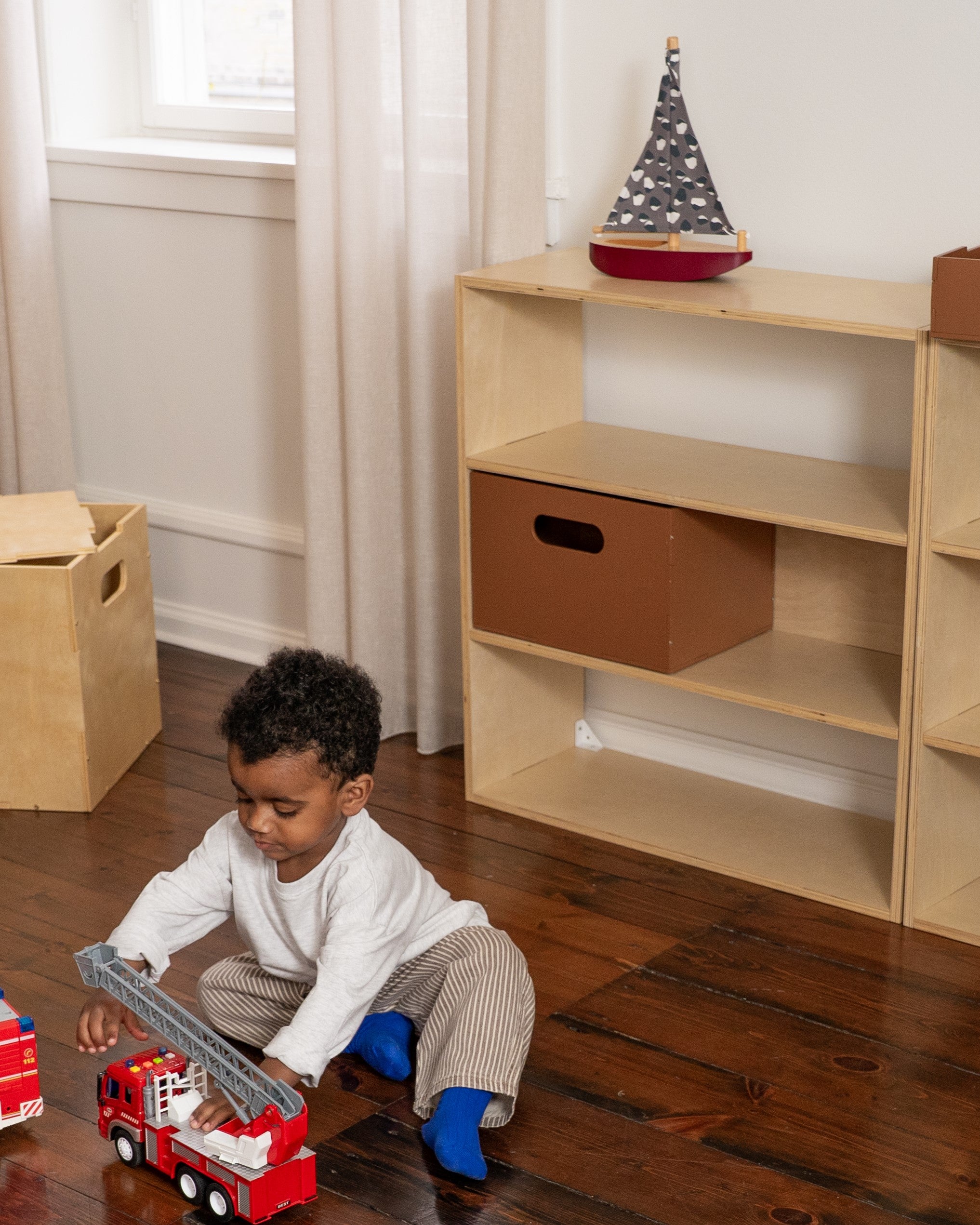 Child playing with a toy fire truck in a room with wooden floors and shelves.