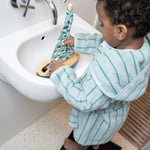 Child in a striped bathrobe standing by a sink holding a green and white toy.