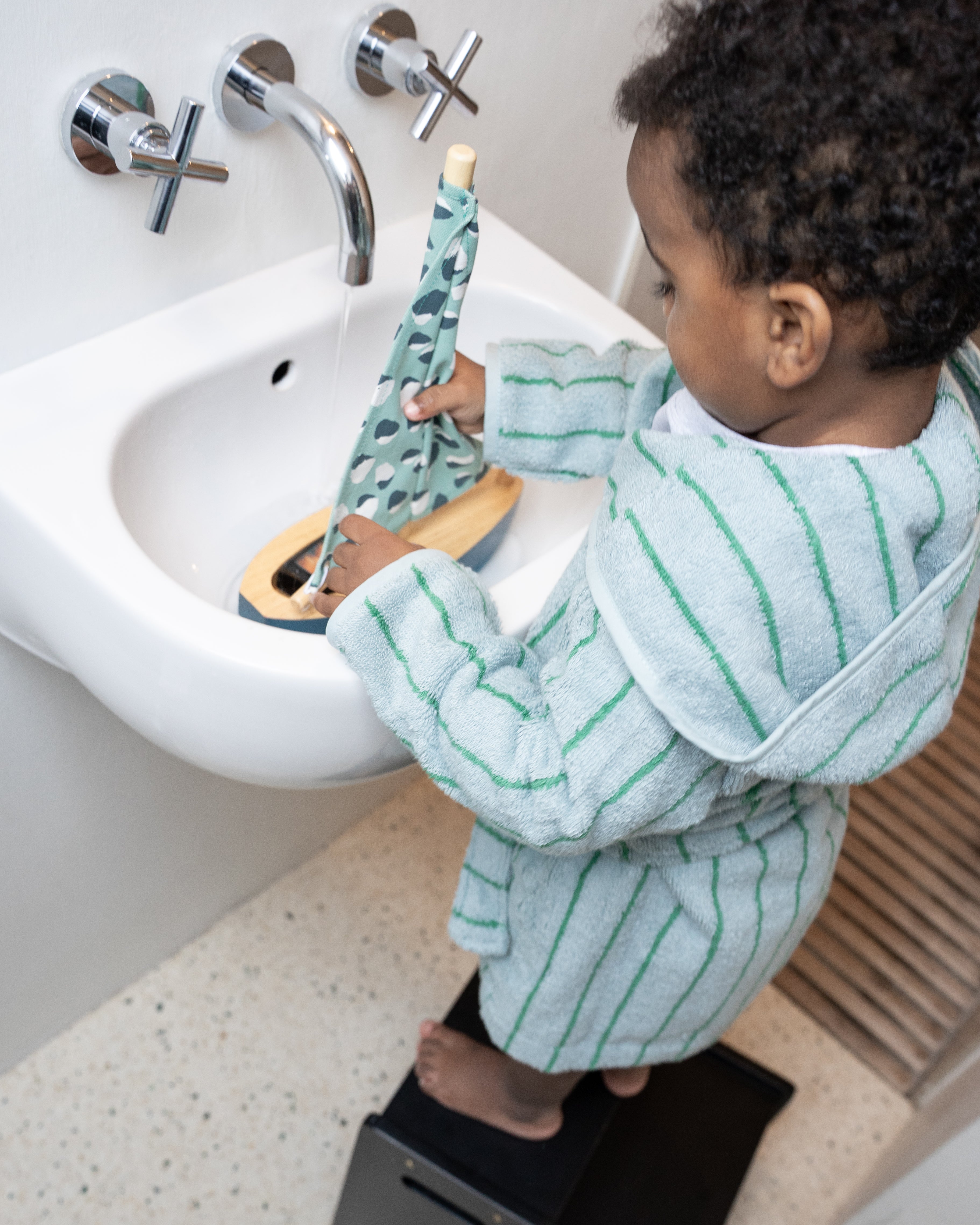 Child in a striped bathrobe standing by a sink holding a green and white toy.