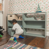 Child playing with a green toy in a room with floral wallpaper and wooden floor.