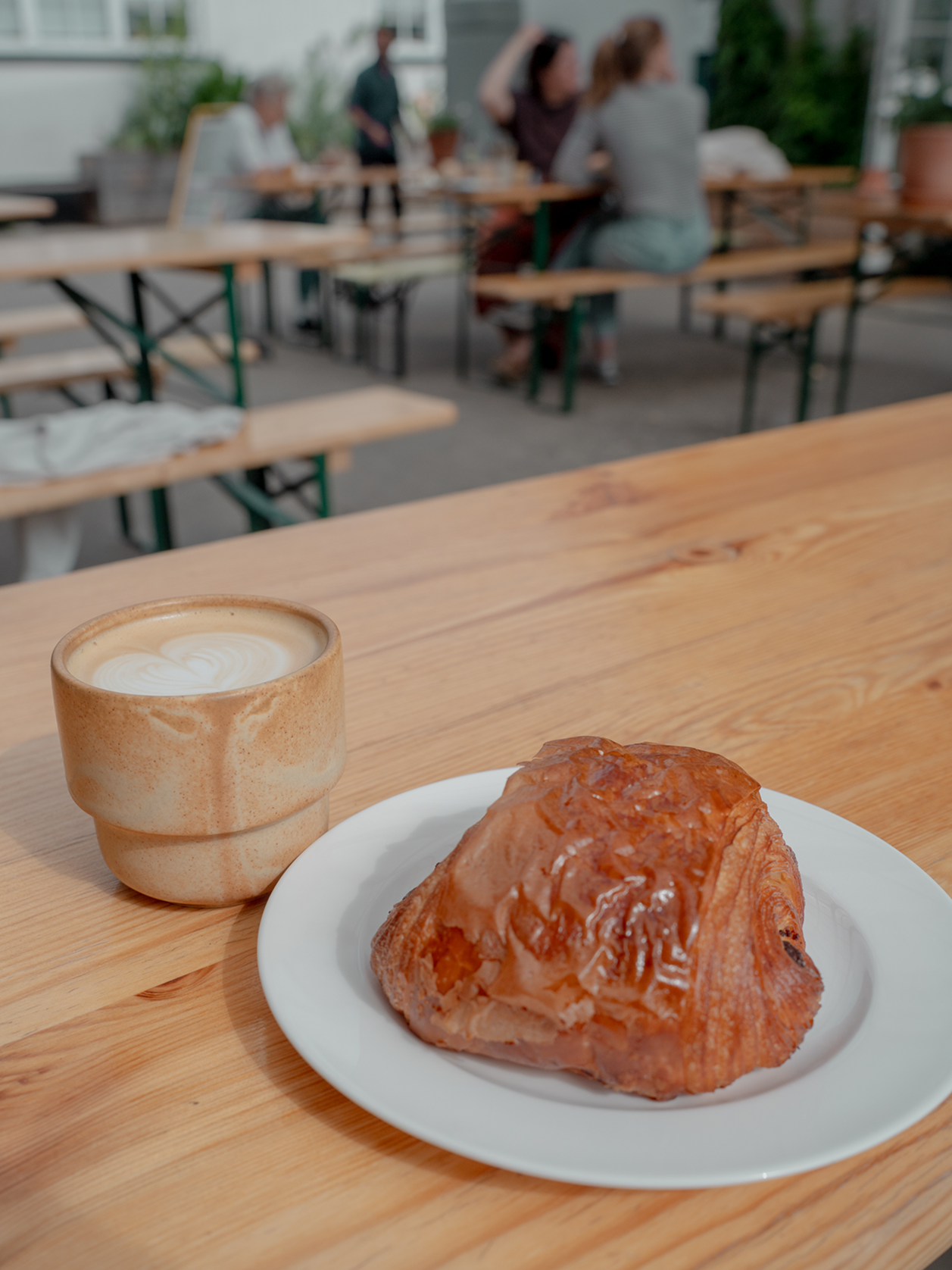 Café au lait in a ceramic cup and a pastry on a plate on a wooden table with people in the background.