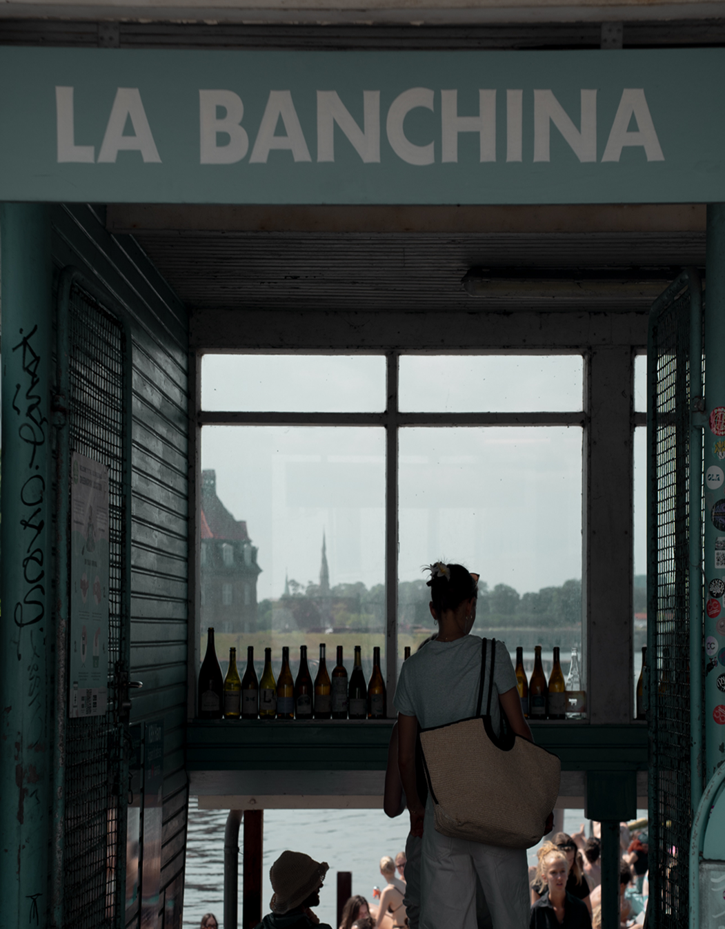 Person standing in front of a window with 'La Banchina' sign, overlooking a waterfront.