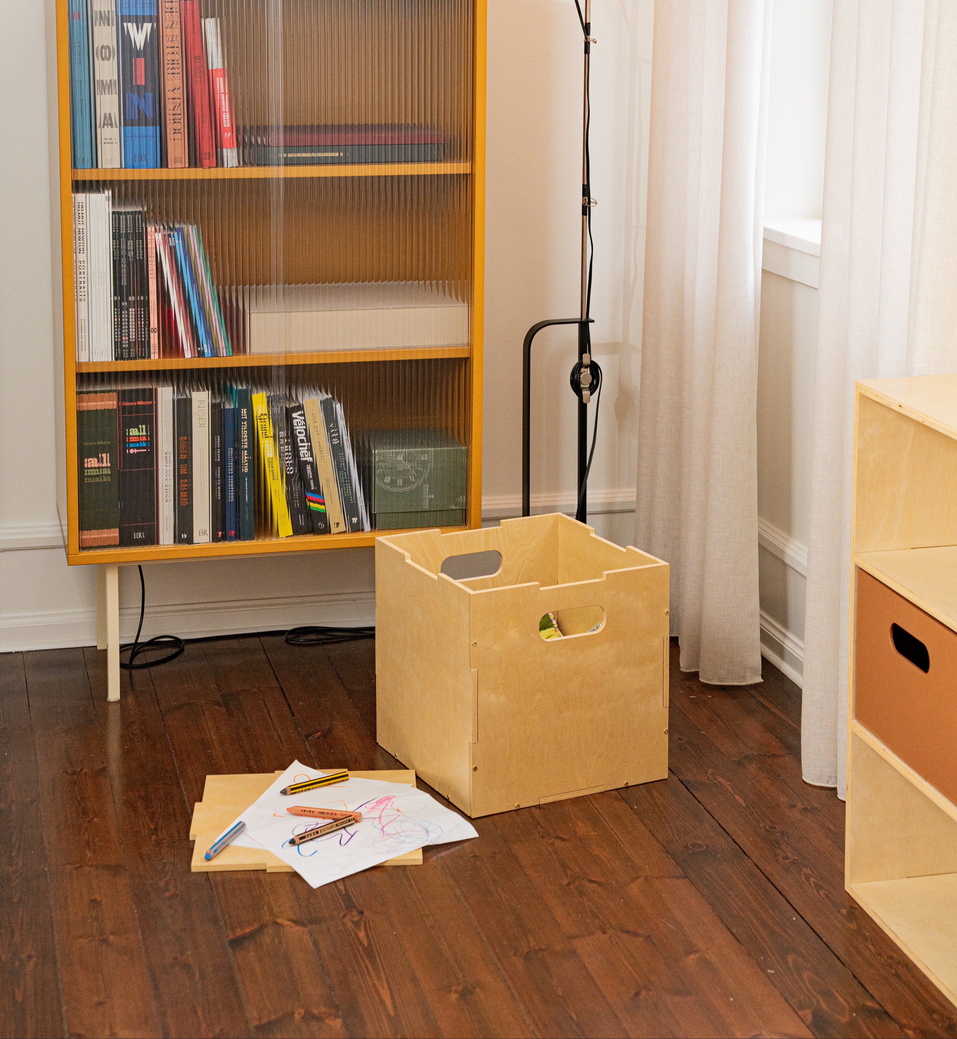 A light natural wood storage bin with integrated handles sits on a dark wood floor in a styled room setting next to a yellow bookshelf and a floor lamp.