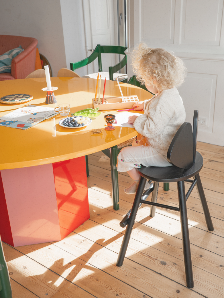 Child sitting at a colorful table with a black chair in a bright room.