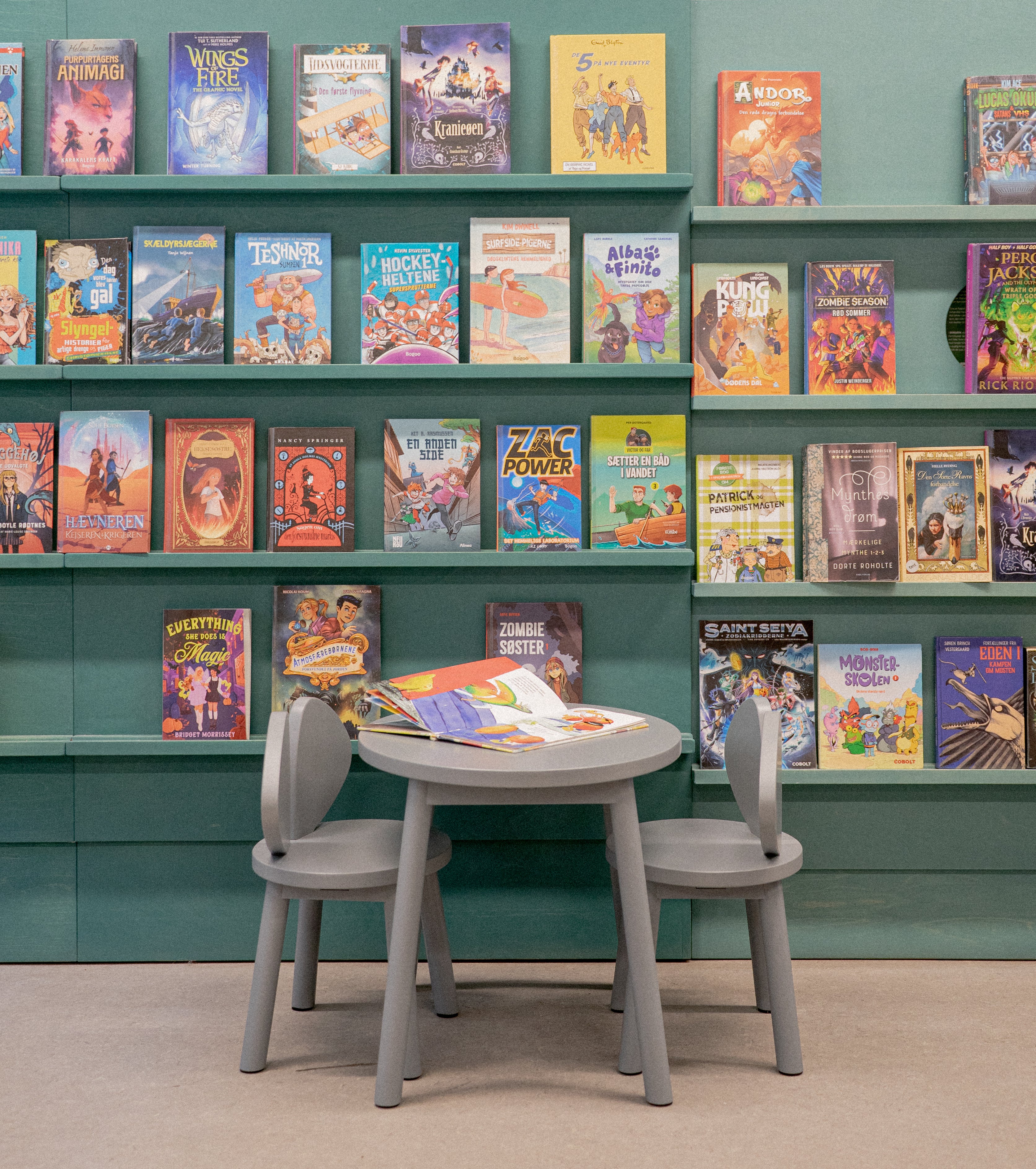 Children's books on shelves with a table and chairs in front of them against a green wall.