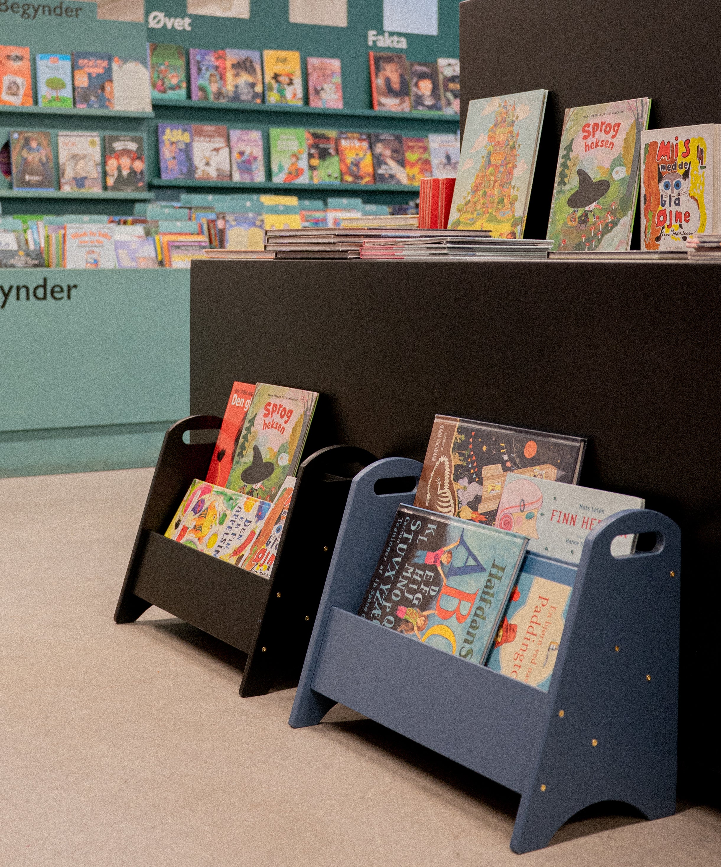Children's books in two stands with a bookshelf of books in the background
