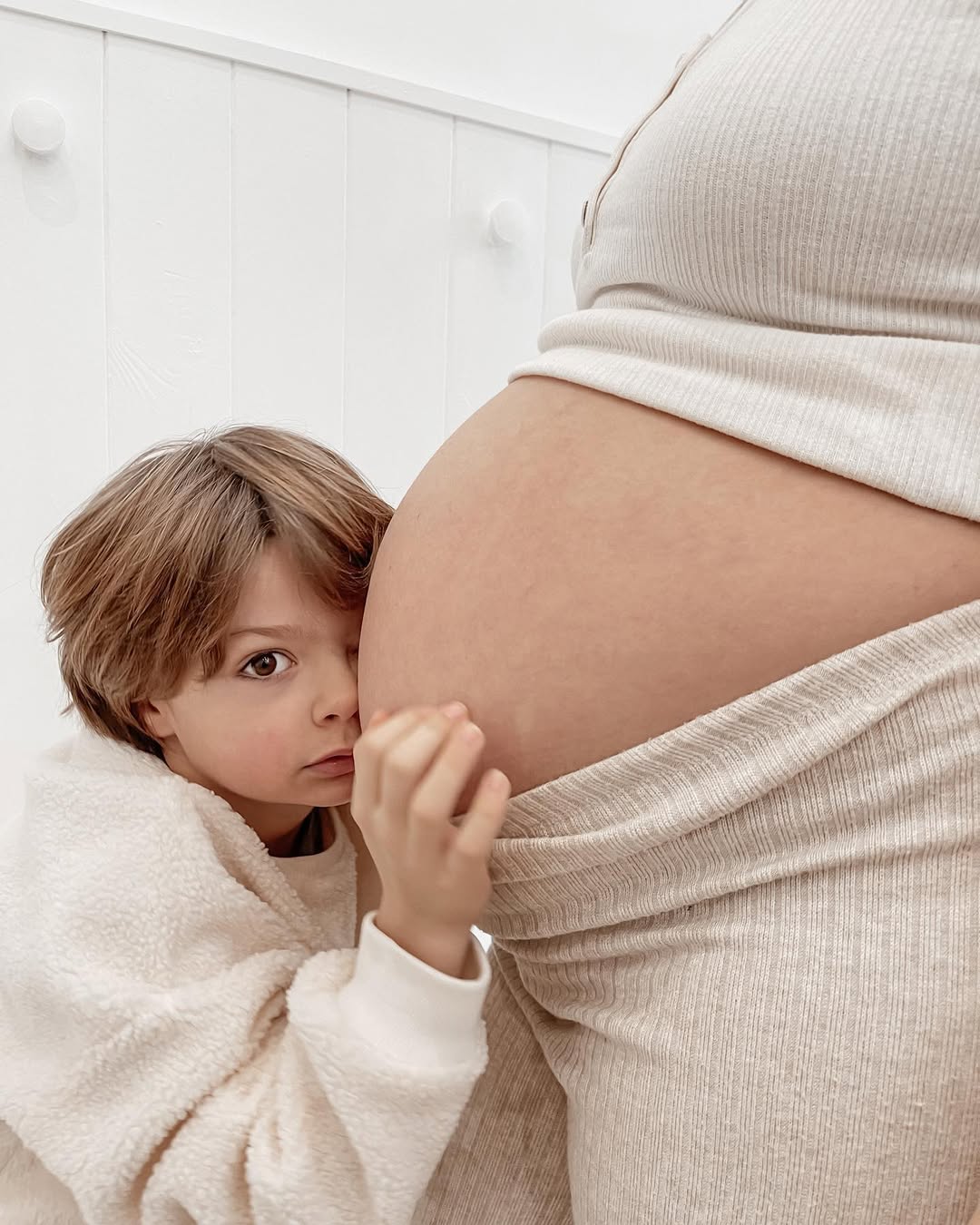 Child touching a pregnant belly, both wearing light-colored clothing against a white background