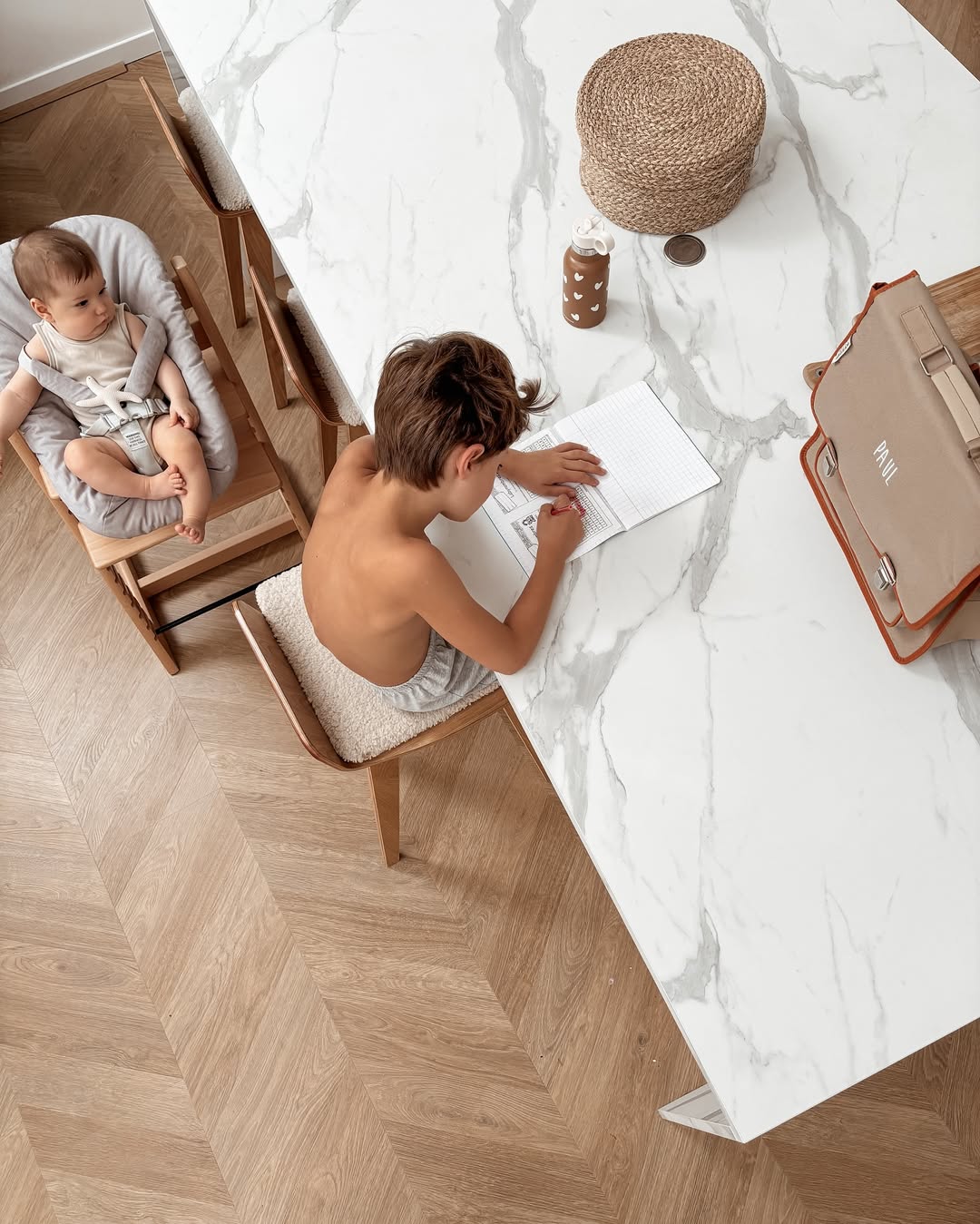 Child sitting at a marble table with a baby on a chair, both using laptops.