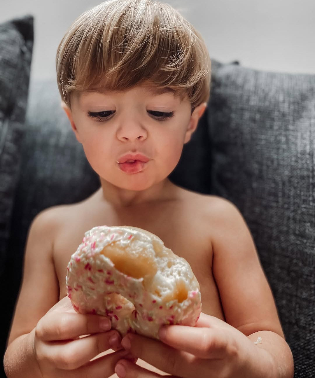 Child holding a cookie with sprinkles and a filling, sitting on a couch.