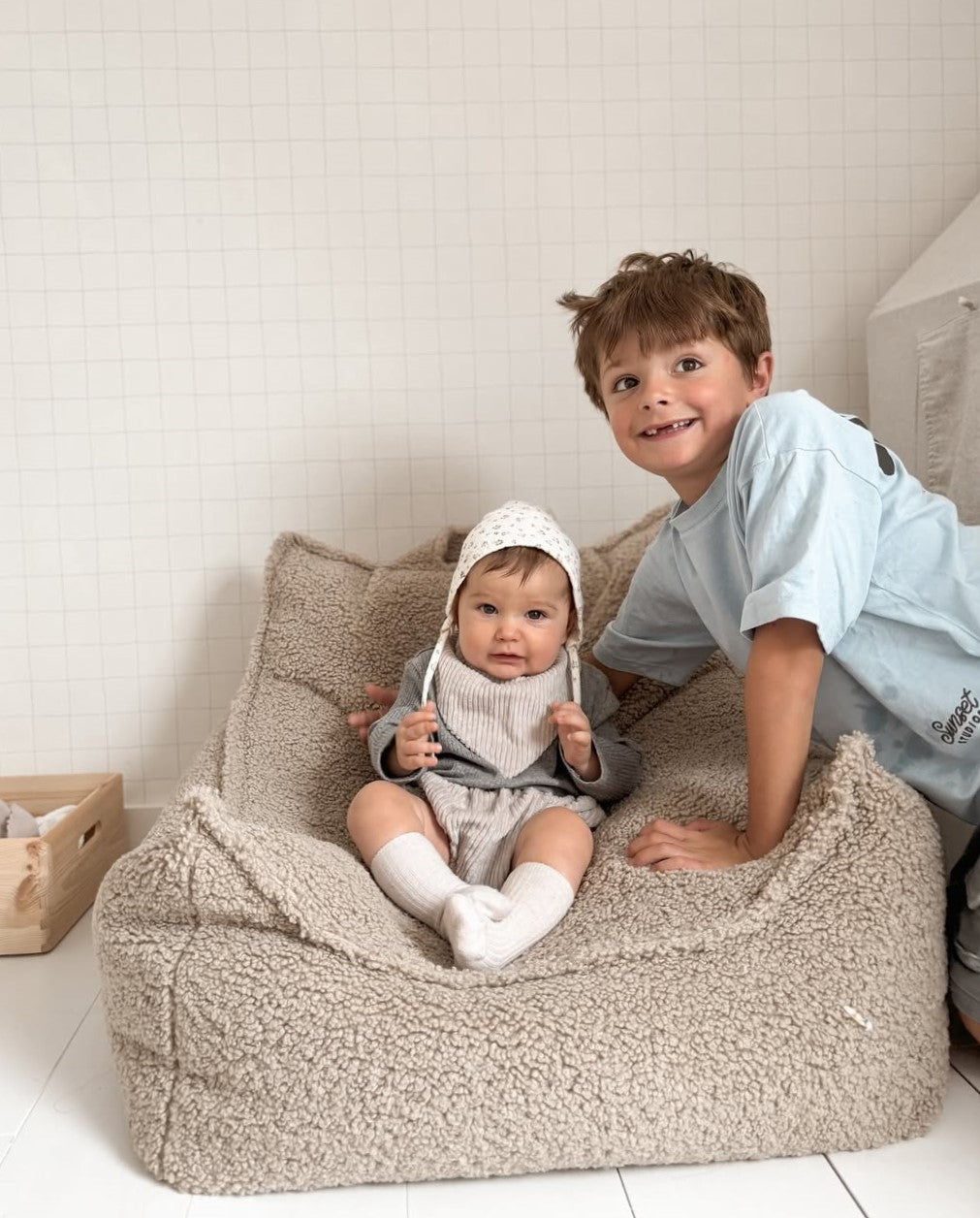Two children on a beige bean bag chair in a room with white walls.