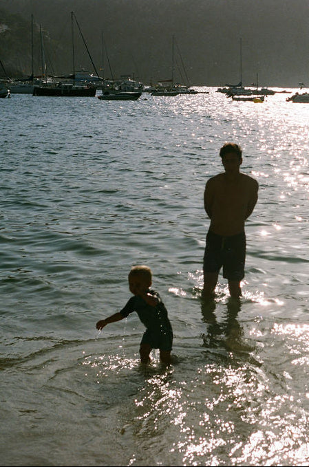 Three people wading in shallow water at a harbor during bright sunlight, with boats and a hillside town visible in the background.