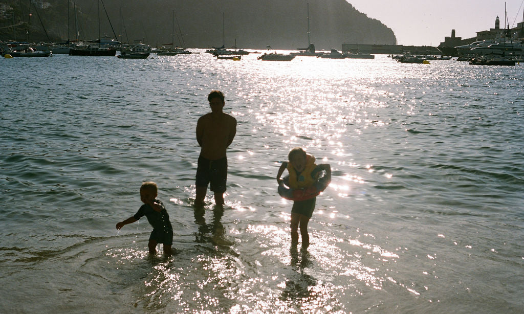 Three people wading in shallow water at a harbor during bright sunlight, with boats and a hillside town visible in the background.