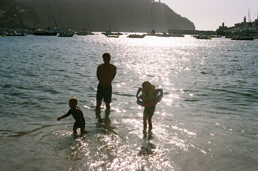 Three people wading in shallow water at a harbor during bright sunlight, with boats and a hillside town visible in the background.