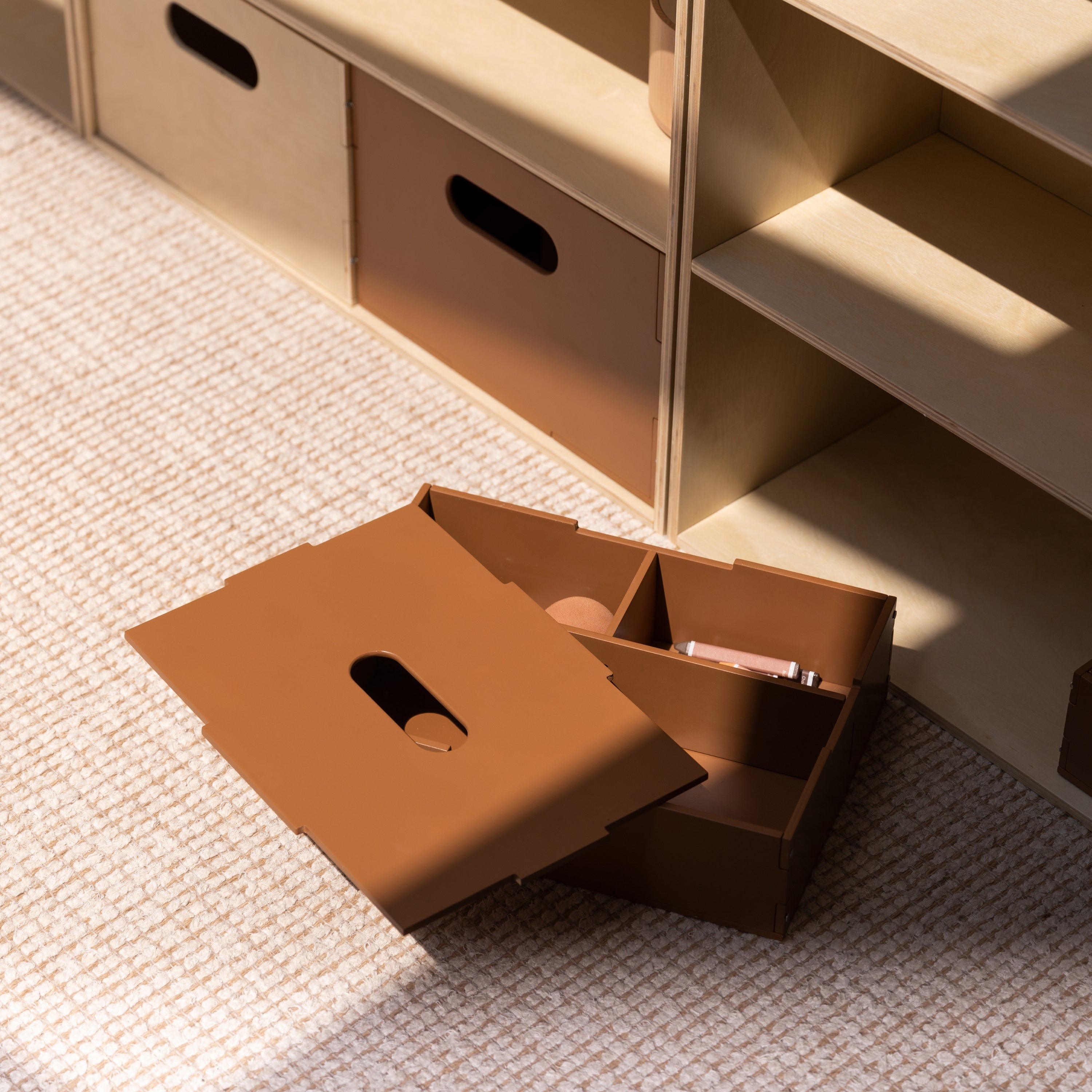 A brown cardboard organizer box with a hinged lid and multiple compartments sits open on a textured beige carpet next to a light wood shelving unit.