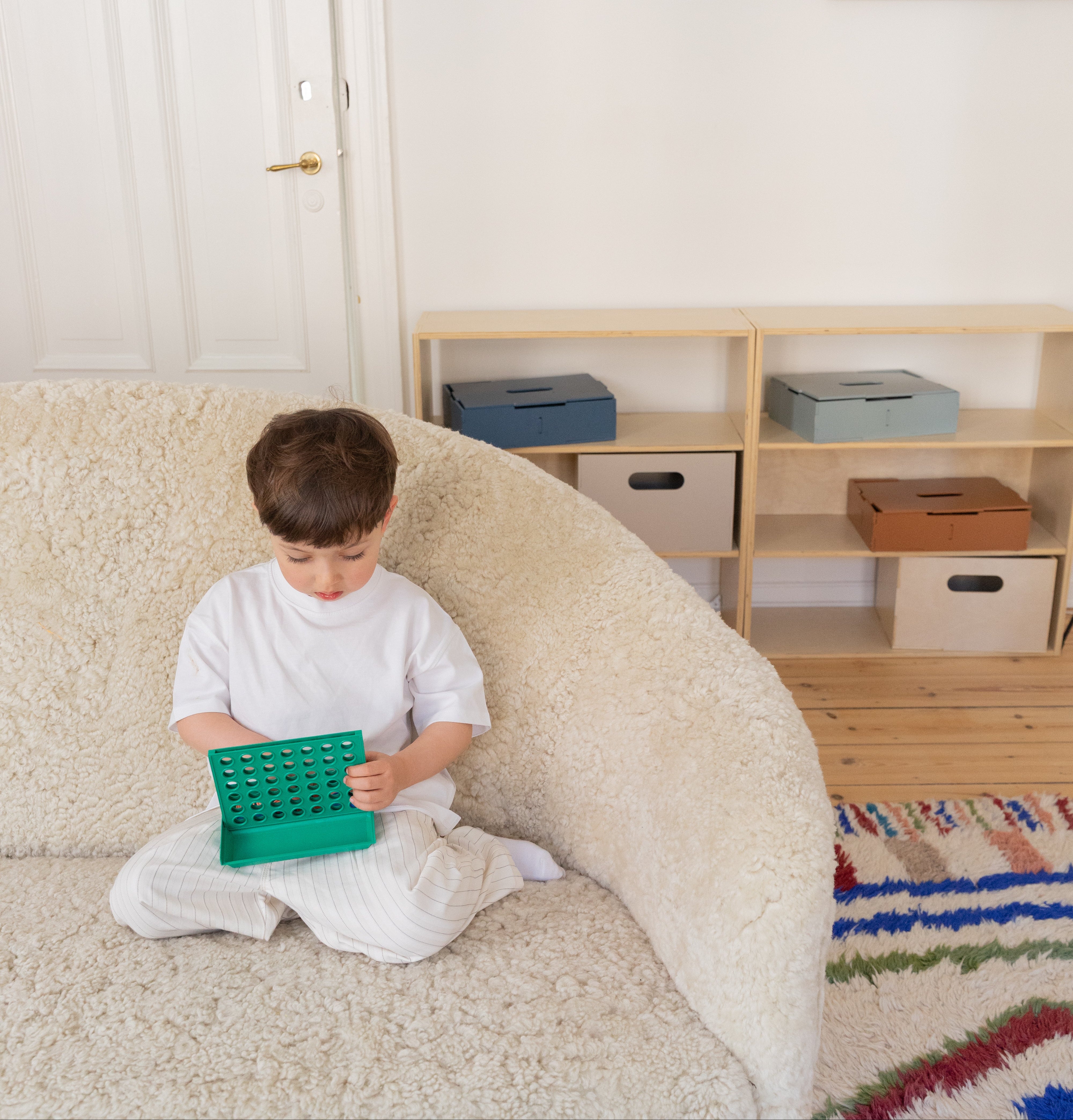 Child sitting on a beige sofa holding a green toy in a room with wooden shelves and a colorful rug.