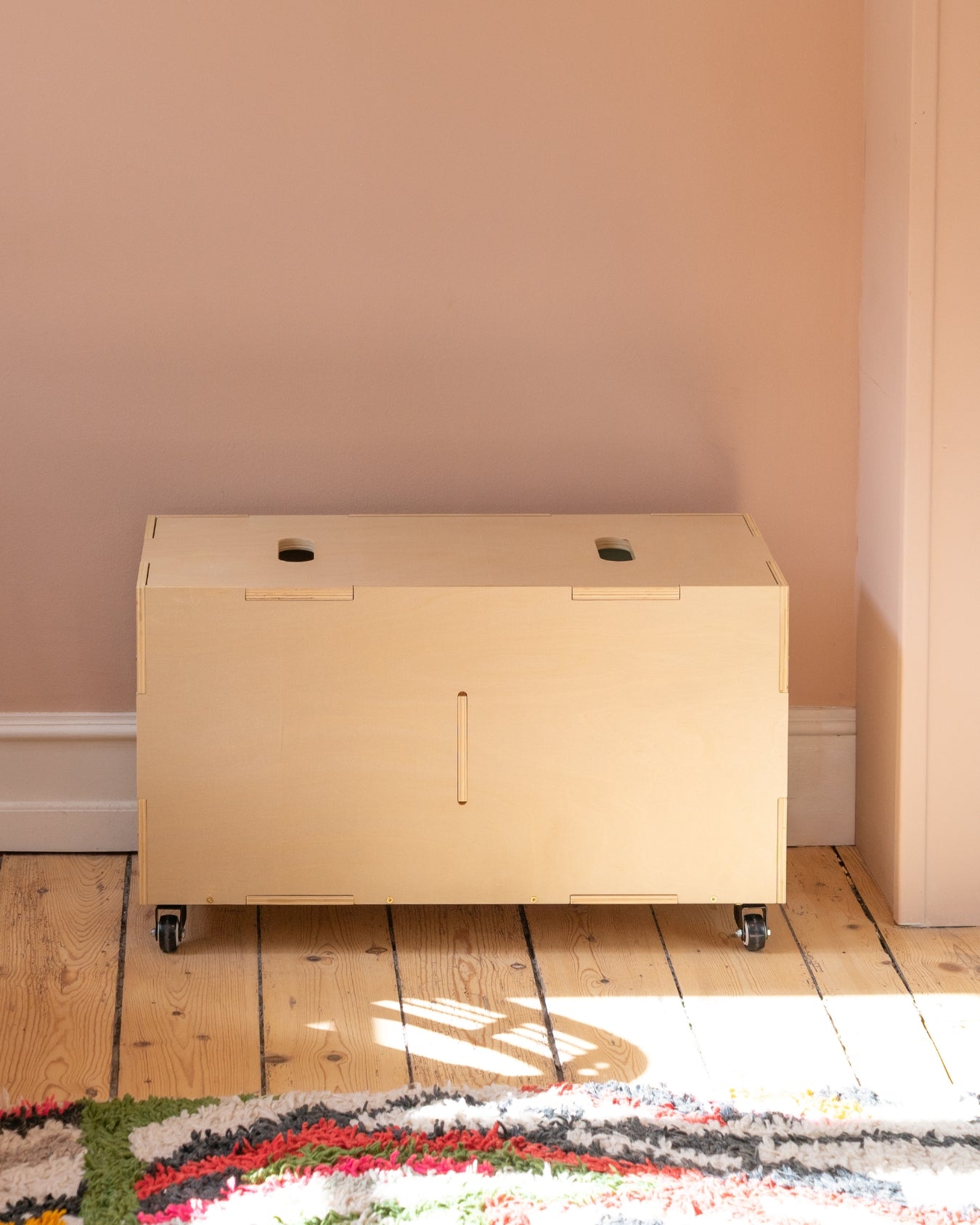 Beige storage cabinet on wheels against a light pink wall with a colorful rug on the floor.