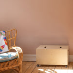 Wicker chair with colorful blanket next to a wooden box on a wooden floor against a beige wall.