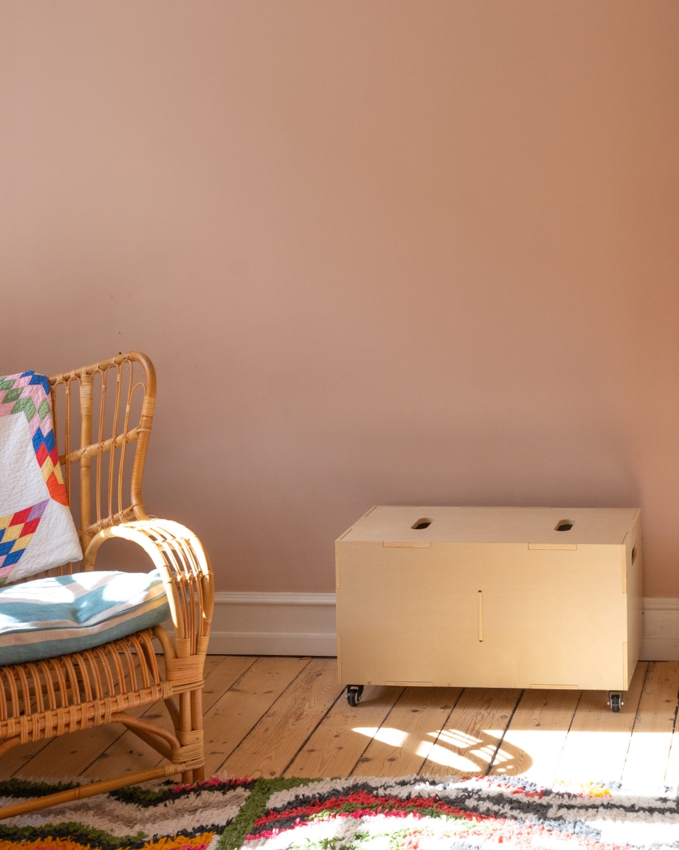 Wicker chair with colorful blanket next to a wooden box on a wooden floor against a beige wall.