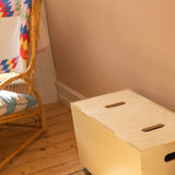 Birch Nofred Cube Long storage bench on wheels, viewed from above beside a wicker chair on a wooden floor.