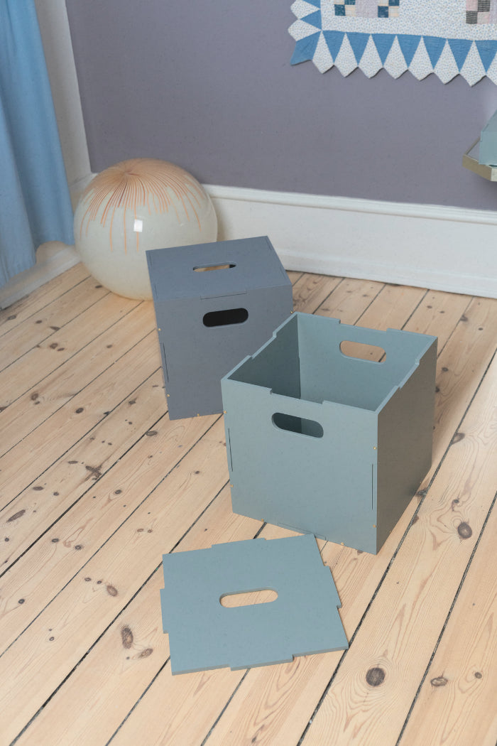 Three gray storage boxes on a wooden floor with a blue wall and decorative elements in the background.