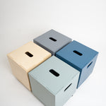 Four minimalist cube storage boxes with handle cutouts, shown in beige, gray, blue, and light green on a clean white background.