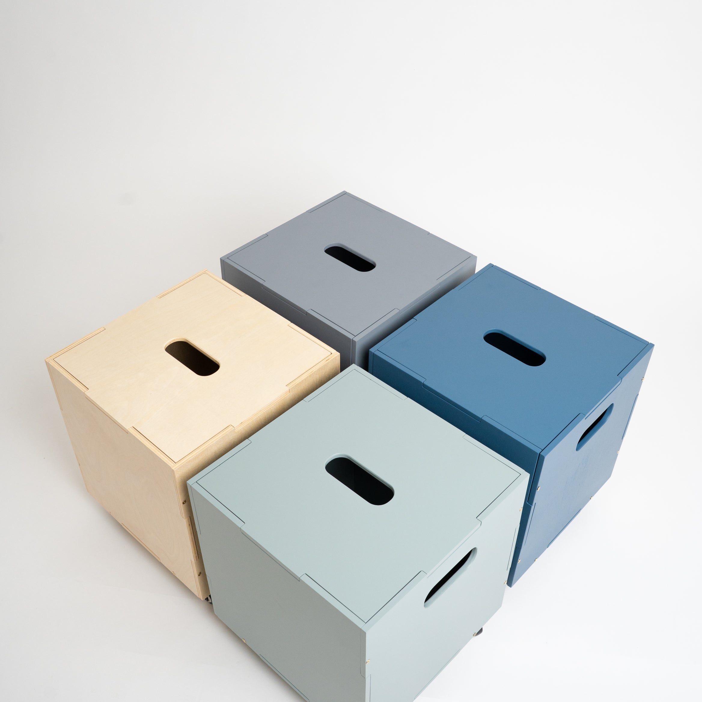 Four minimalist cube storage boxes with handle cutouts, shown in beige, gray, blue, and light green on a clean white background.