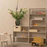 A modern modular shelving unit in a warm beige tone, styled with a large green vase of flowers, wooden storage boxes, colorful children's toys, and a small yellow step stool in a minimalist room.